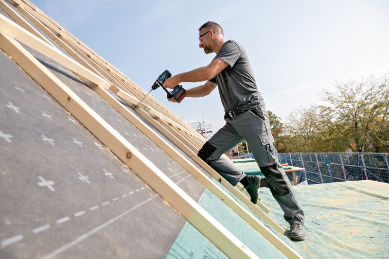 En person iført sikkerhedsudstyr bruger en batteridrevet boremaskine til at fastgøre træ på et skrånende tag.